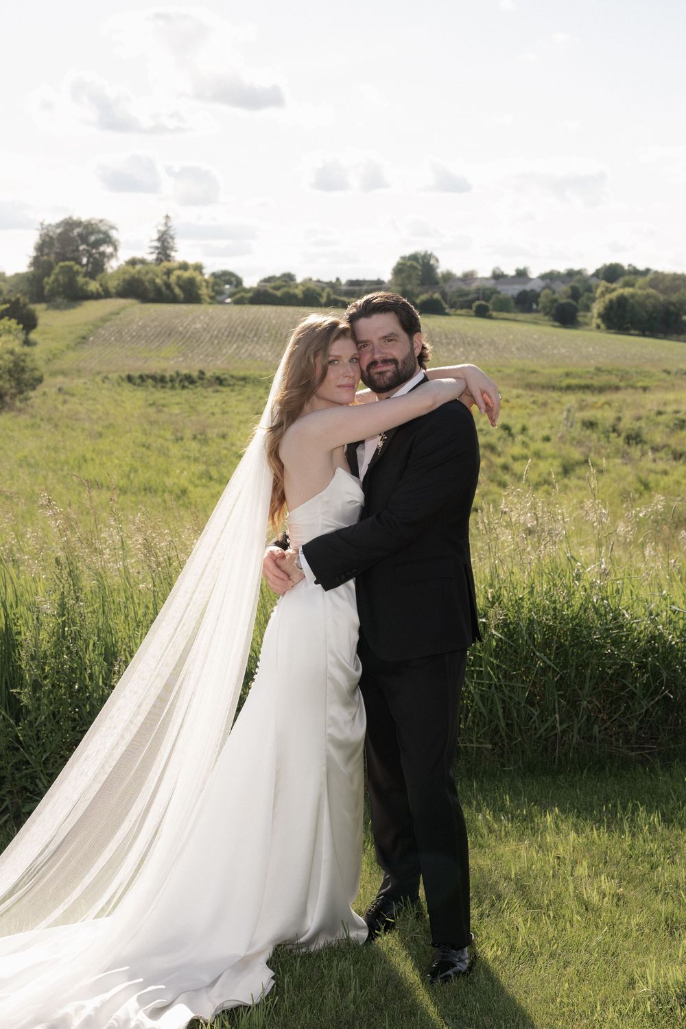 Bride and groom embracing in a lush green field, bride wearing a flowing white gown with long veil on their wedding day.