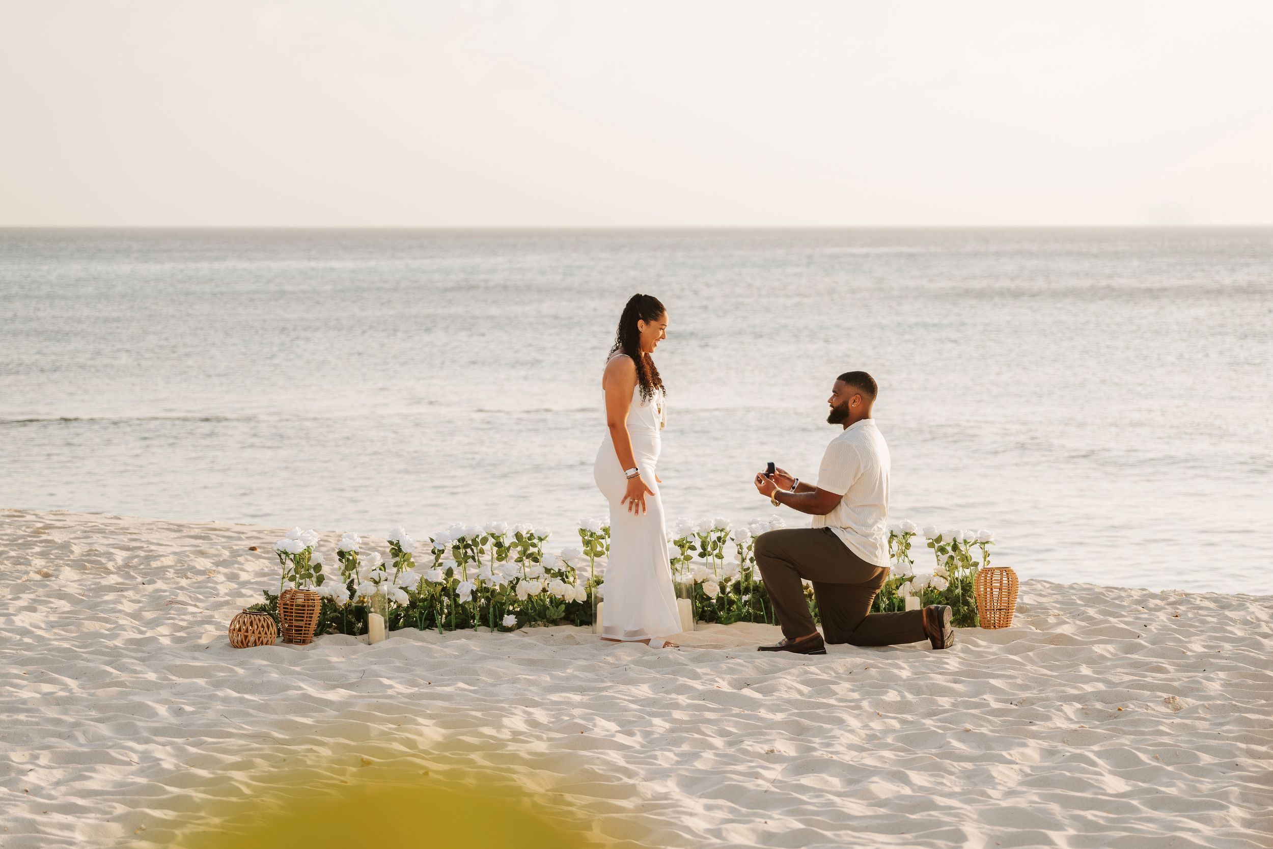 Couple holding hands at sunset beach proposal setup with floral arch, candles, and Marry Me sign on sandy shore.