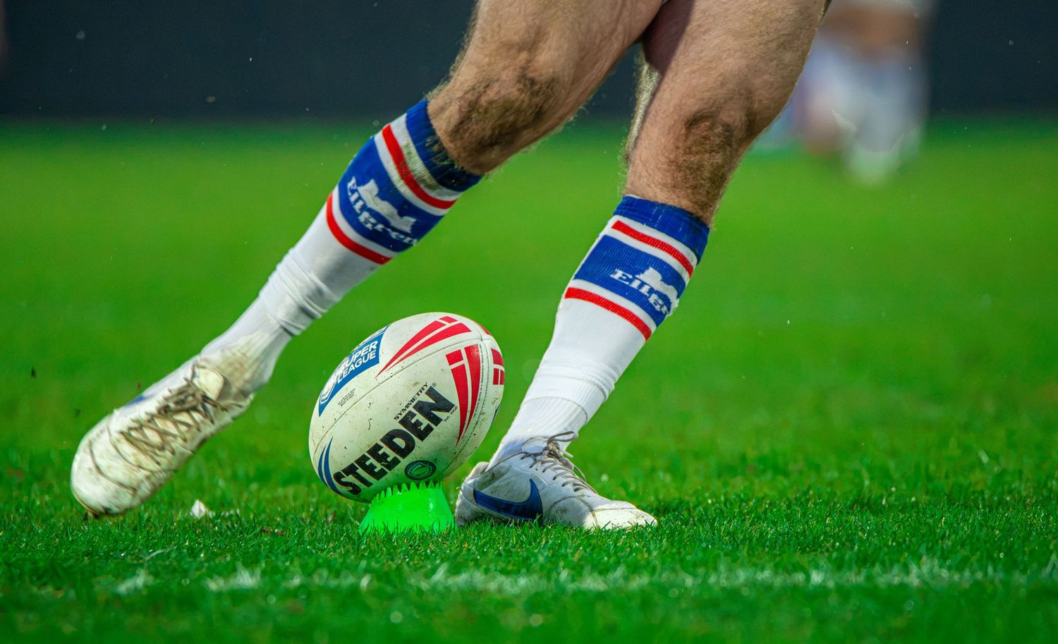 Close-up of rugby league player placing a Steeden match ball on a kicking tee on grass.