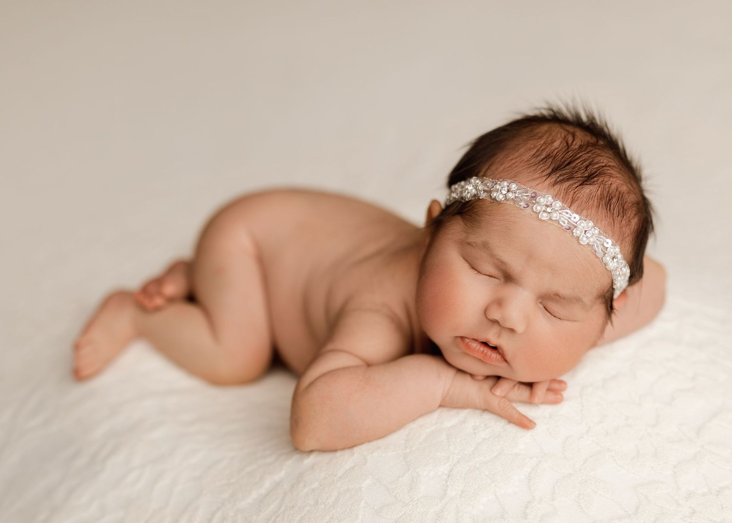 Sleeping newborn baby girl wearing a delicate rhinestone headband, posed on a soft white fluffy blanket.