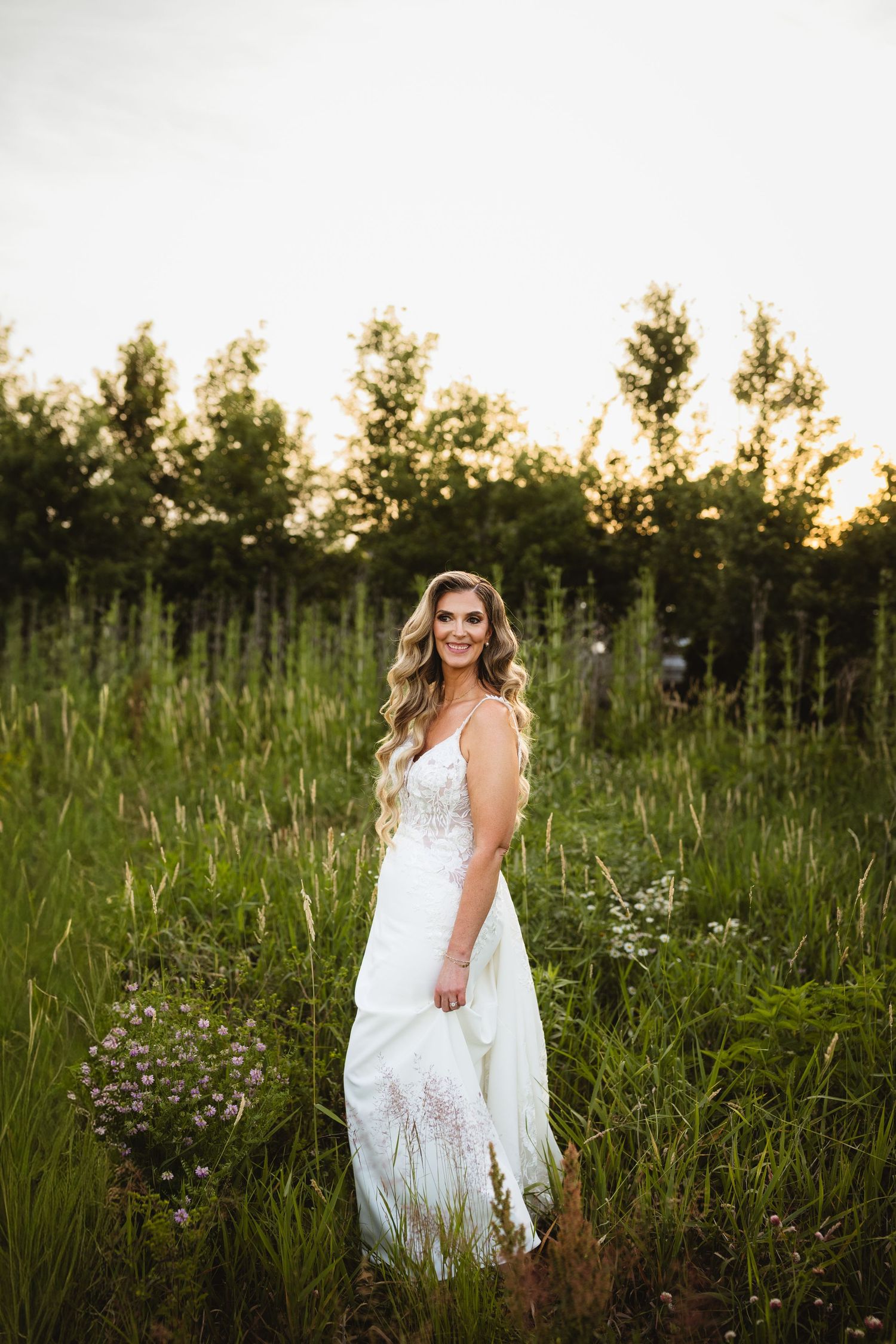 Smiling bride in white lace gown standing in lush green meadow with wildflowers at golden hour.