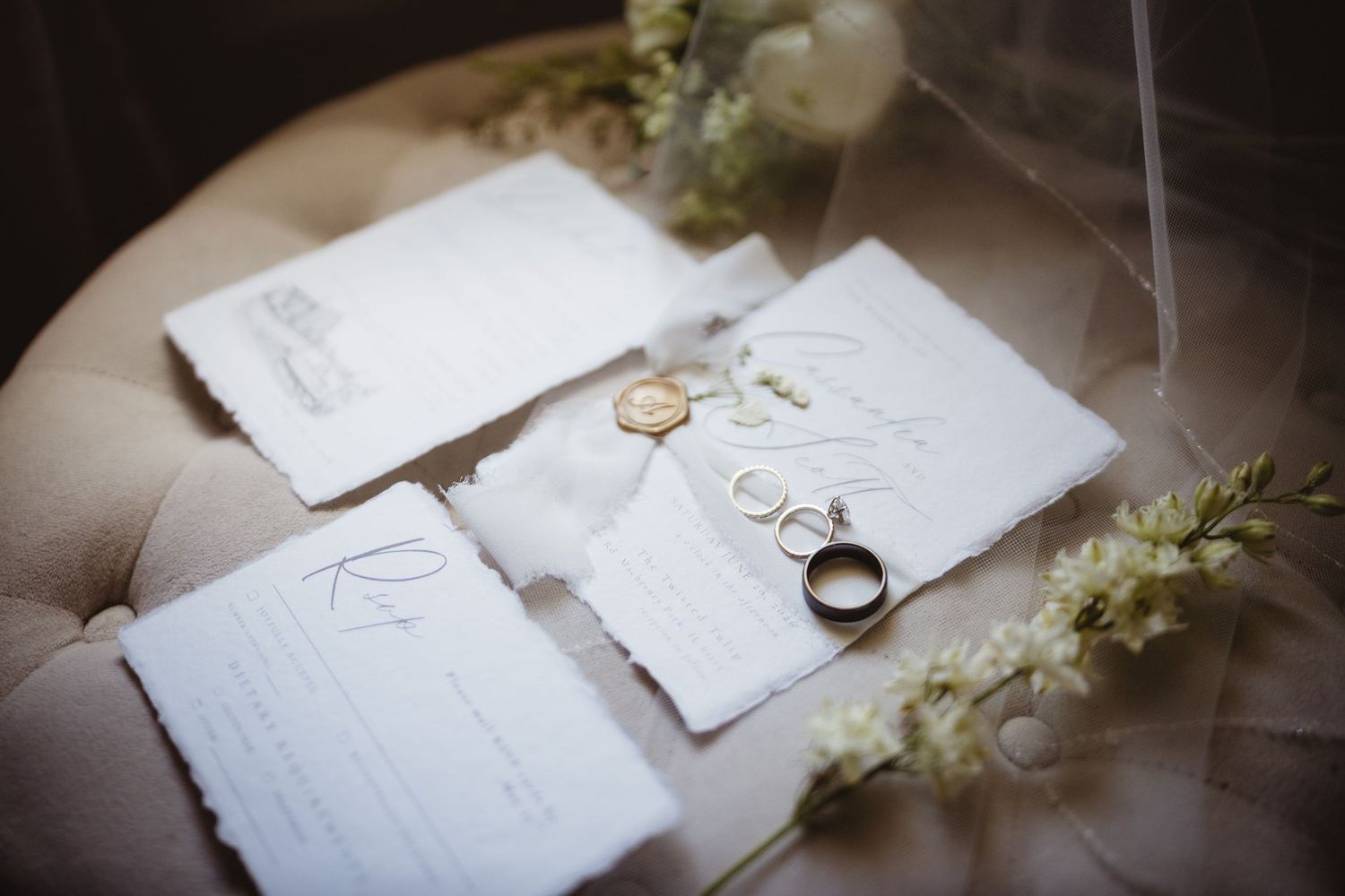 Elegant wedding stationery with gold wax seal, rings, and delicate white flowers on a tufted cream surface.