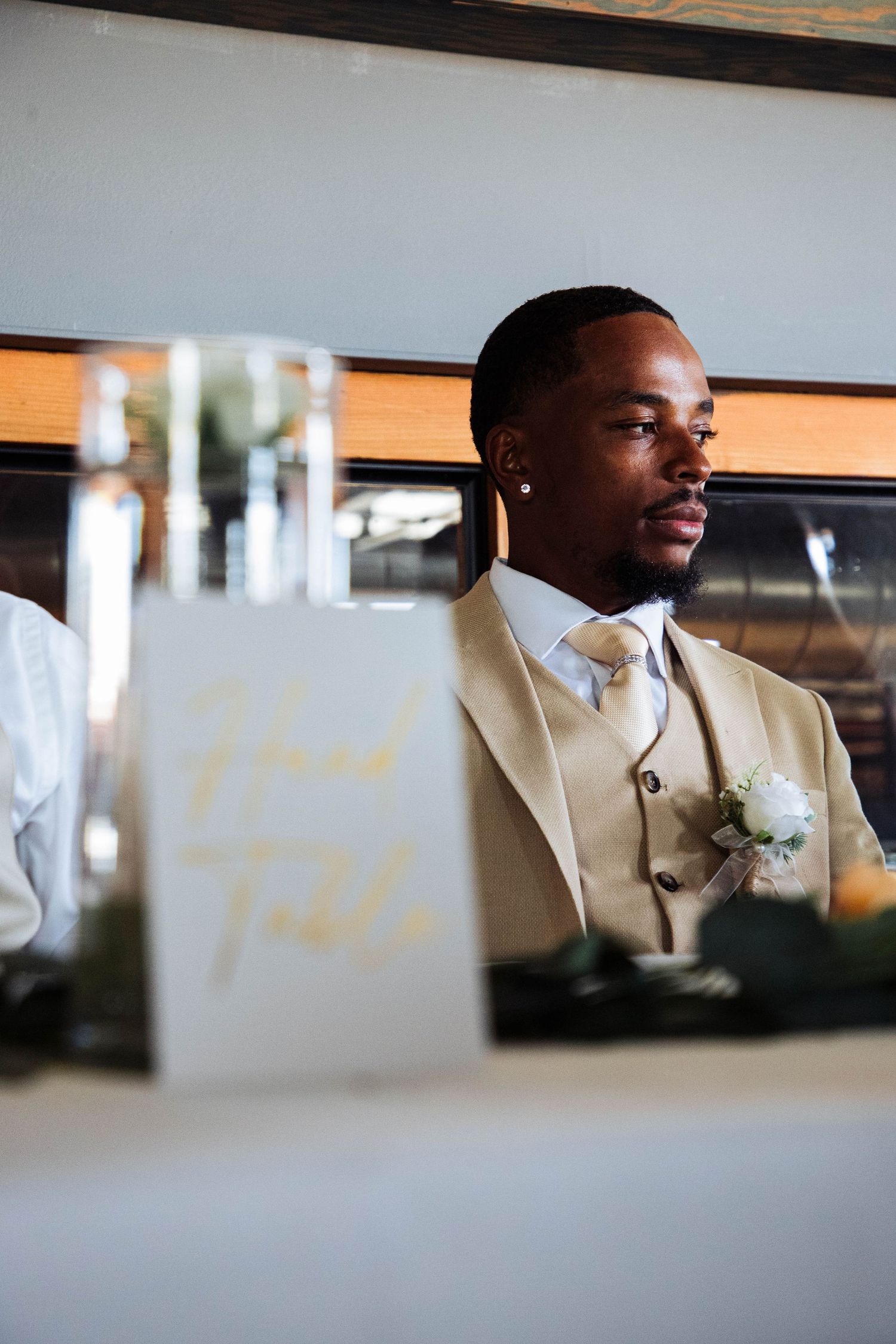 Groom in beige suit with boutonniere looking contemplative at wedding reception.