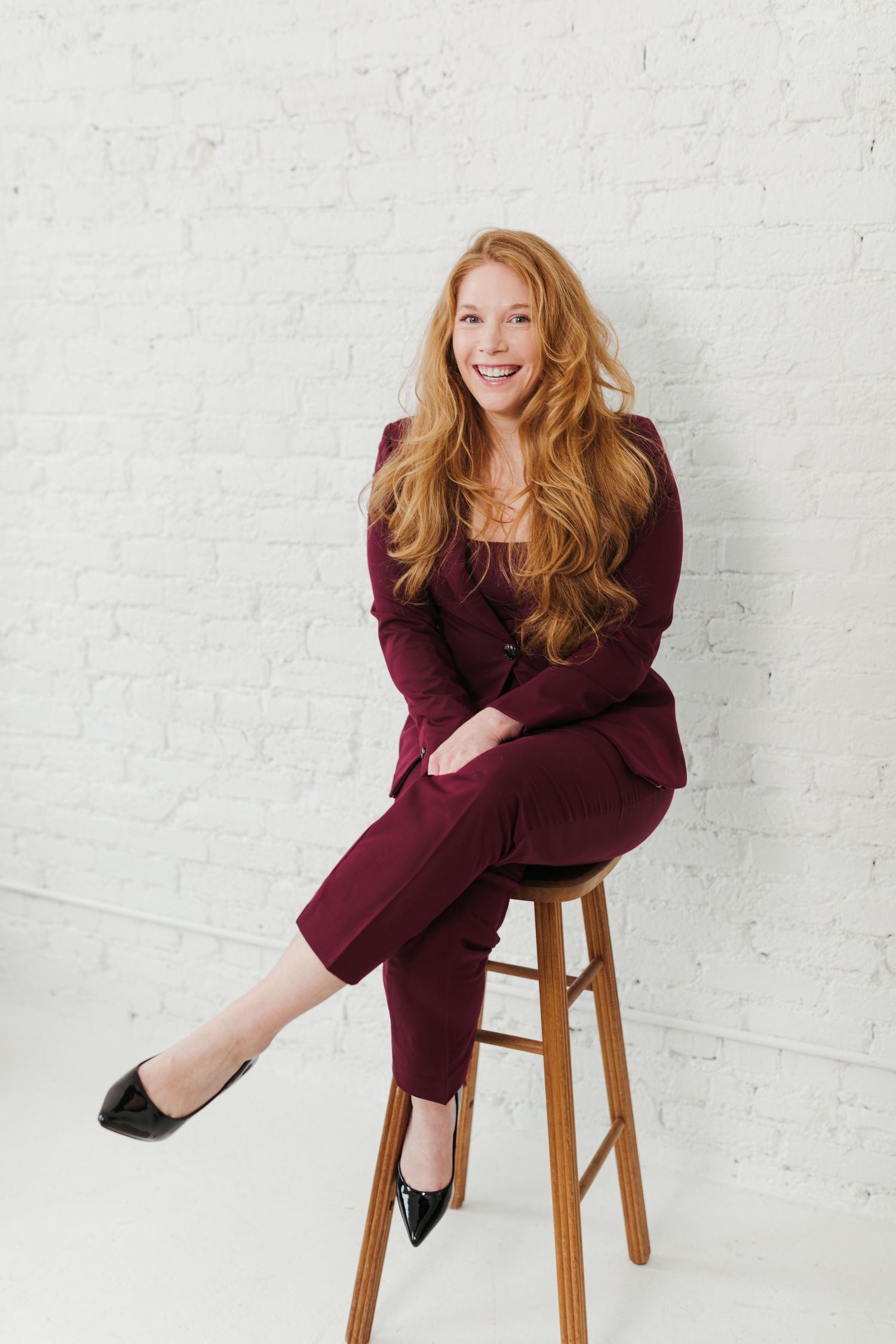 A smiling redhead woman in a burgundy pantsuit sits on a wooden stool against a white brick wall.