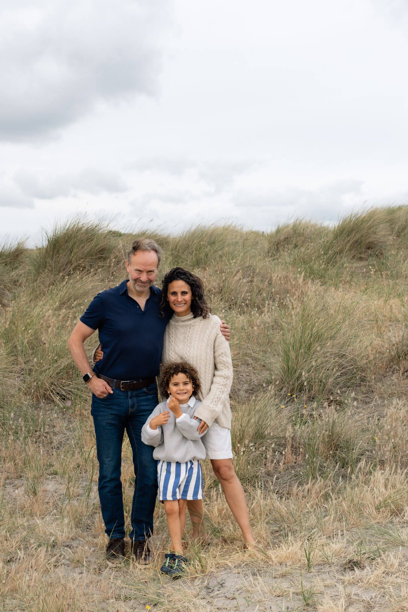 A family of three posing together on a sandy beach dune with tall grass and cloudy skies behind them during a family photoshoot in Sussex