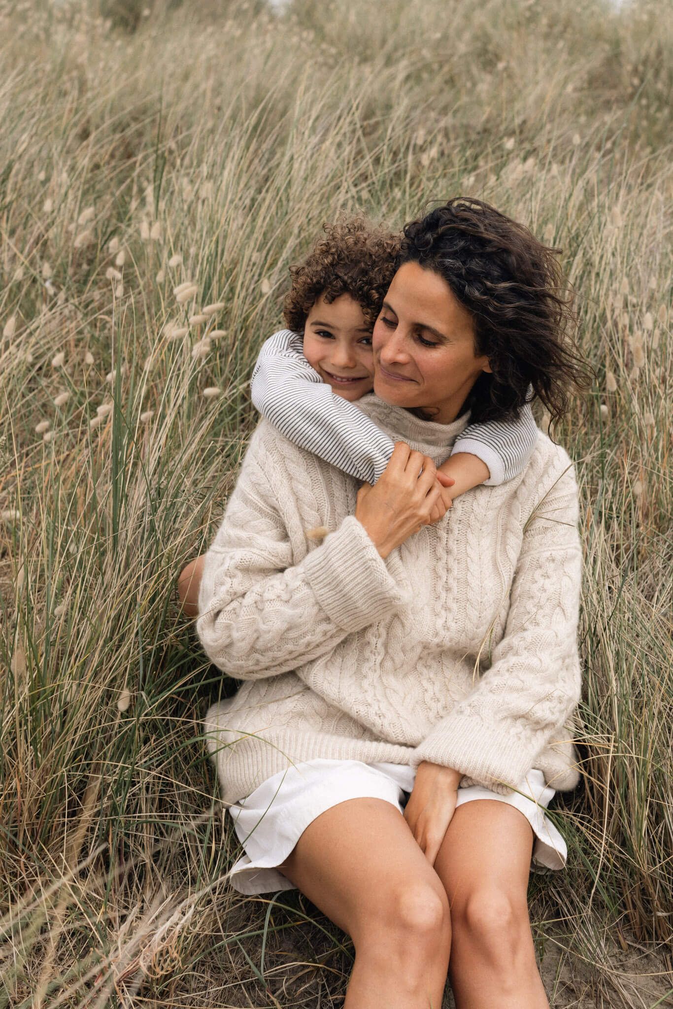 A mother and child share a warm embrace sitting in tall grass, both wearing cozy knit sweaters during an outdoor family photoshoot in Sussex