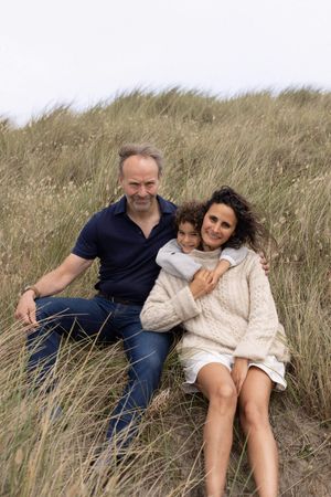 A family of three sits together in coastal grass dunes, smiling warmly, during a family photography session in Sussex
