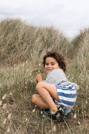 Smiling curly-haired boy sitting in tall beach grass during a family photography session in Sussex