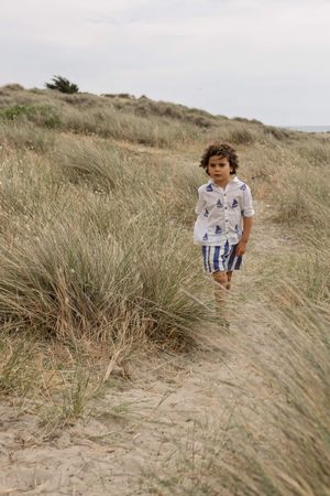 A young boy in a sailboat-print shirt and striped shorts walks along a sandy beach path through coastal dunes in Sussex