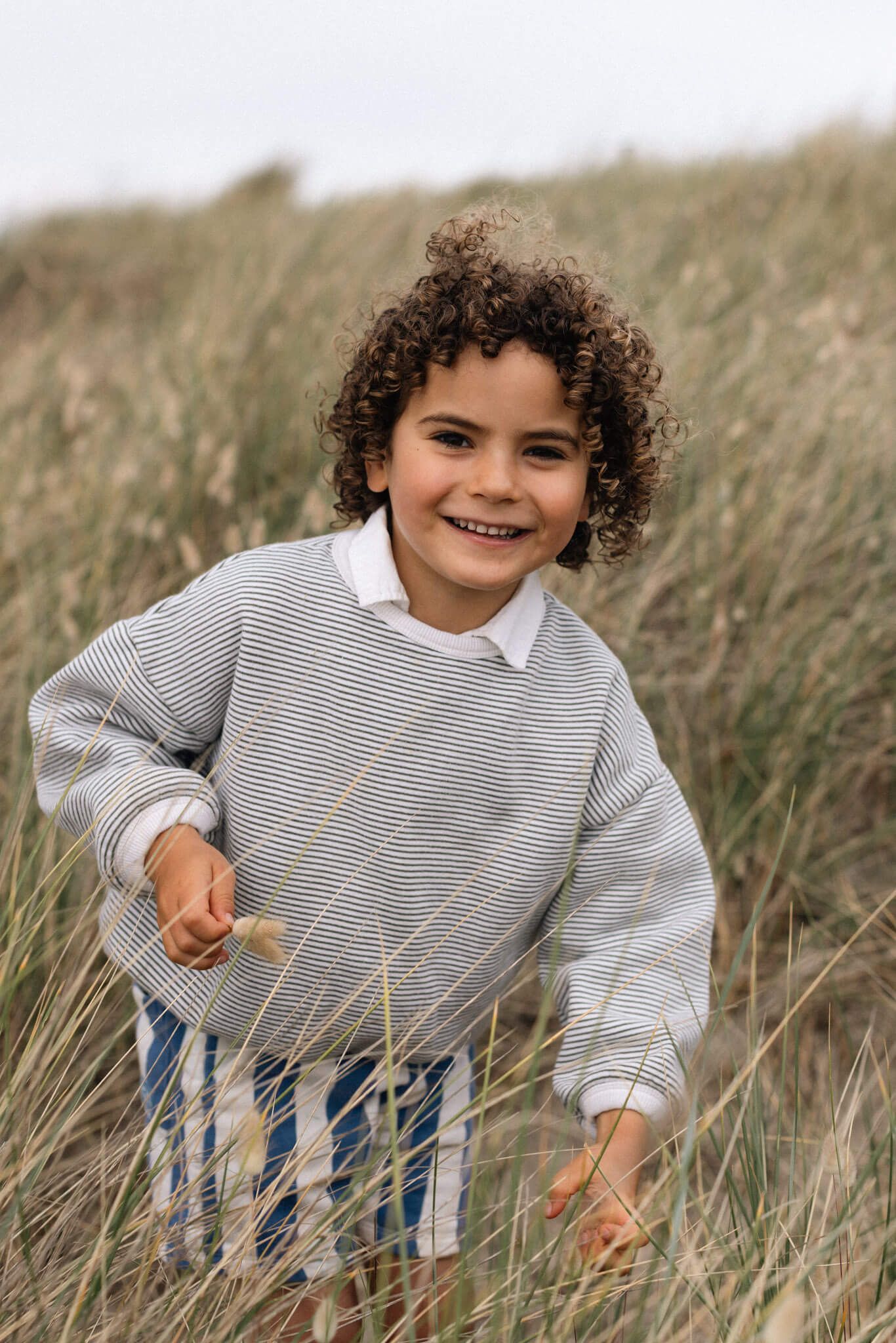 A smiling child with curly hair wearing a striped sweatshirt plays among tall grass on a beach dune in Sussex
