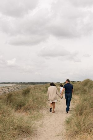 Family walking along a sandy beach path through grass dunes on a cloudy day during a family portrait session in Sussex