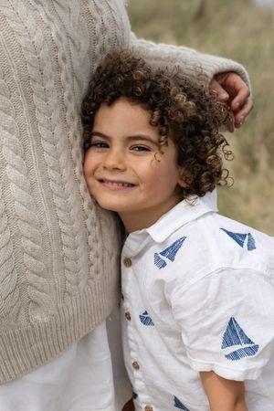 Curly-haired boy smiling and leaning against adult in beige knit sweater during a family beach photography session in Sussex