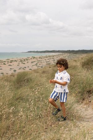 Young boy in nautical shirt and striped shorts walking through beach dunes on a cloudy day during a family portrait session in Sussex