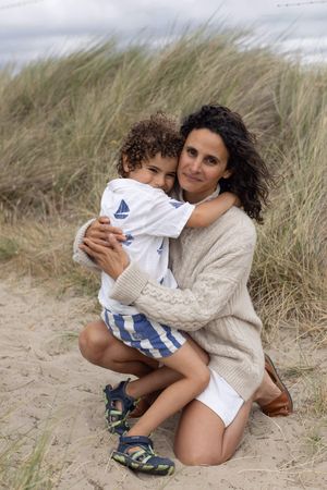 A mother hugs her young curly-haired son on a sandy beach dune with tall grass in the background during a family portrait session in Sussex