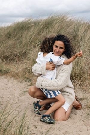 A woman with curly hair hugs a young child on a sandy beach with tall grass and cloudy skies during a family portrait session in Sussex