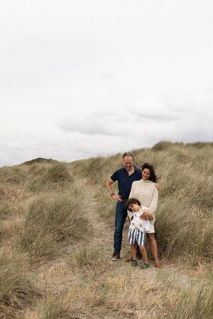 Family of three posing on sandy grass dunes under cloudy sky during a family portrait session in Sussex