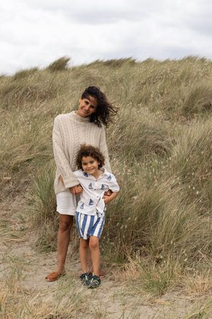 A woman and young child pose together on sandy beach dunes with tall grass, wearing casual summer clothing on a cloudy day.