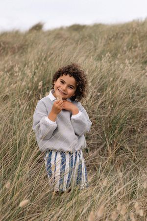 Smiling child with curly hair wearing a grey sweater and striped pants standing in tall beach grass in Sussex