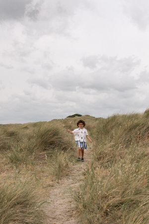 A young boy with curly hair runs along a sandy coastal path through tall beach grass under a cloudy sky during a family photography session
