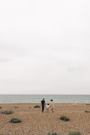 Family of three walking on a pebble beach toward the calm sea on an overcast day during a family portrait session in Sussex