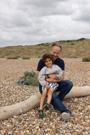 Father and son sitting on driftwood log on pebble beach during a family portrait session in Sussex
