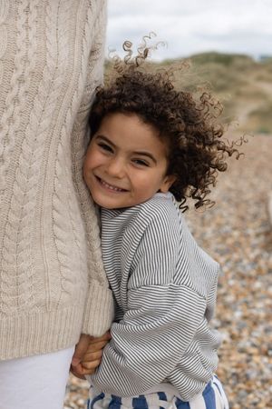 A smiling curly-haired boy hugging his mothers leg on a pebble beach during a family portrait session in Sussex