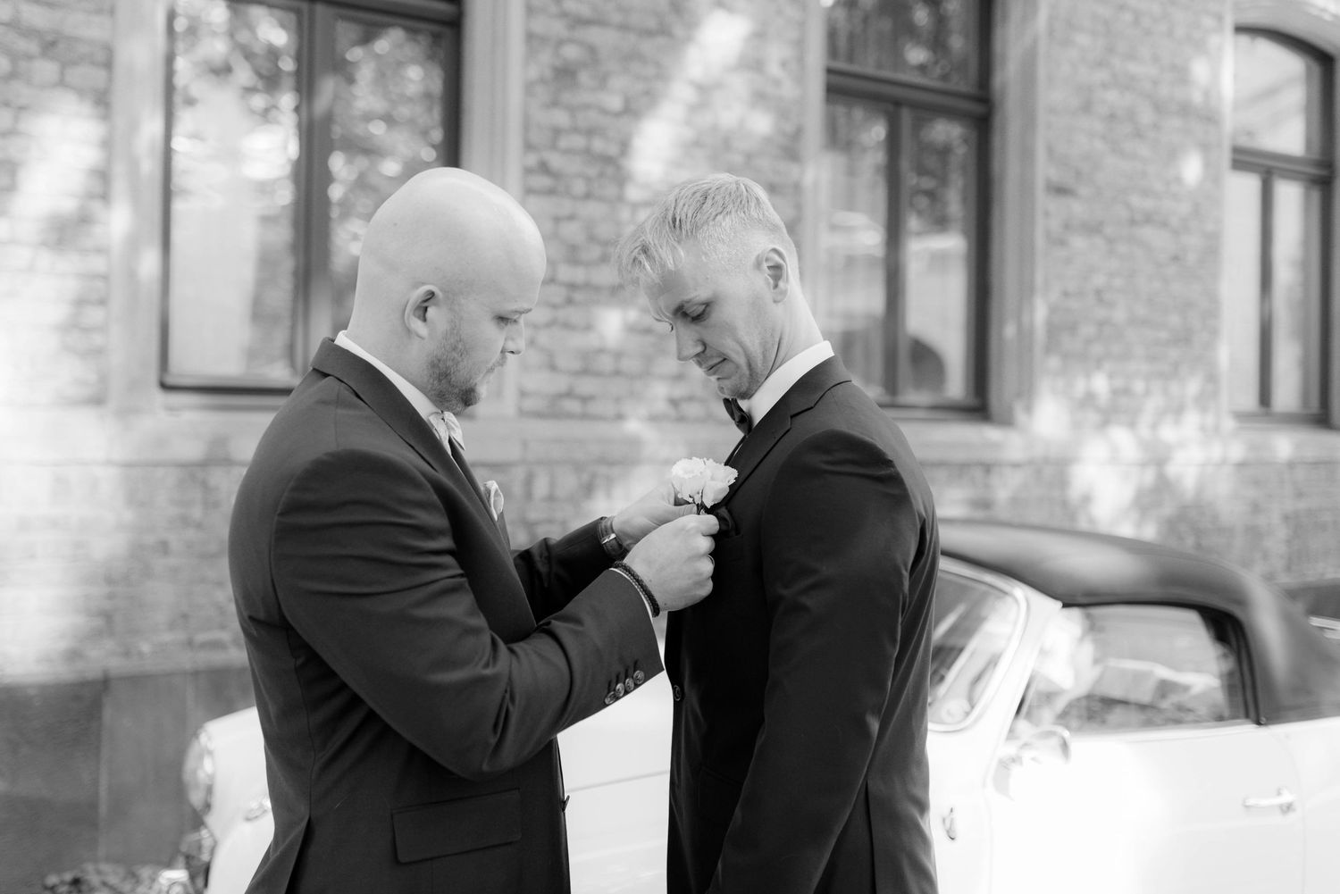 Two grooms in black suits share a tender moment as one pins a boutonniere on the other outdoors.