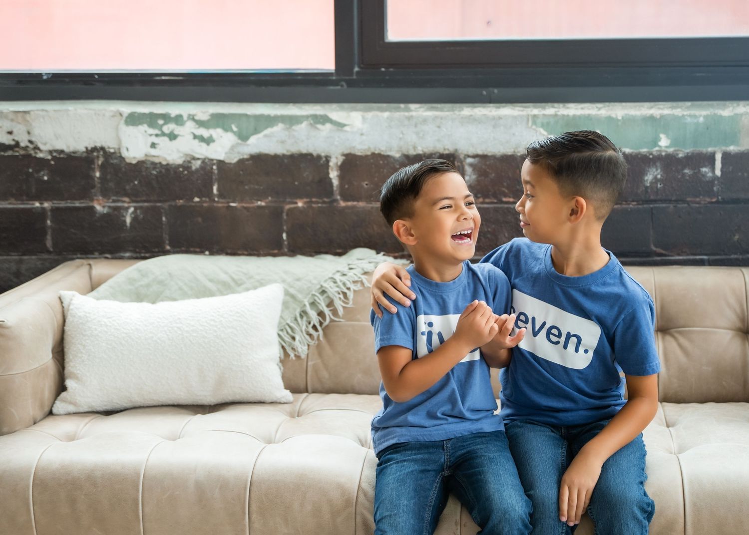 Two young boys in matching blue shirts laugh together on a beige couch in a modern interior setting.