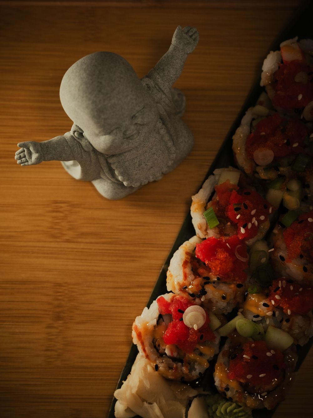 Stone Buddha figurine posed beside a colorful sushi platter with sesame seeds and toppings on a wooden surface.