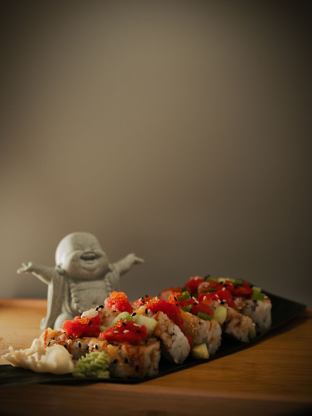 Sushi rolls topped with red salsa on slate board beside smiling Buddha figurine on wooden table.