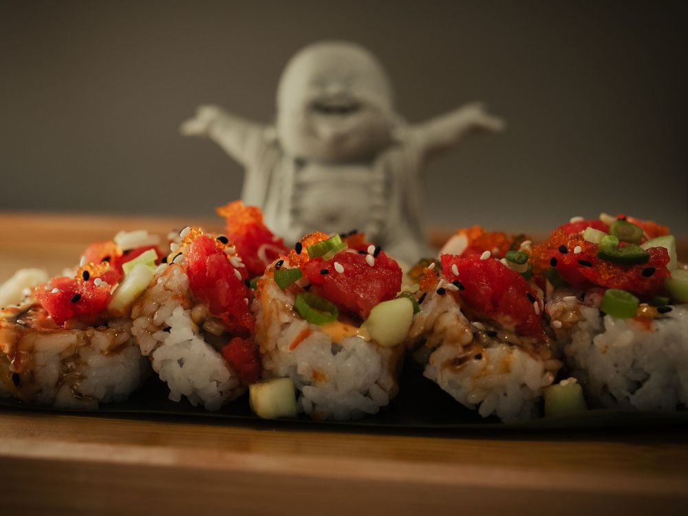 Sushi rolls topped with red pepper, green onions, and sesame seeds on a dark plate with a Buddha statue.