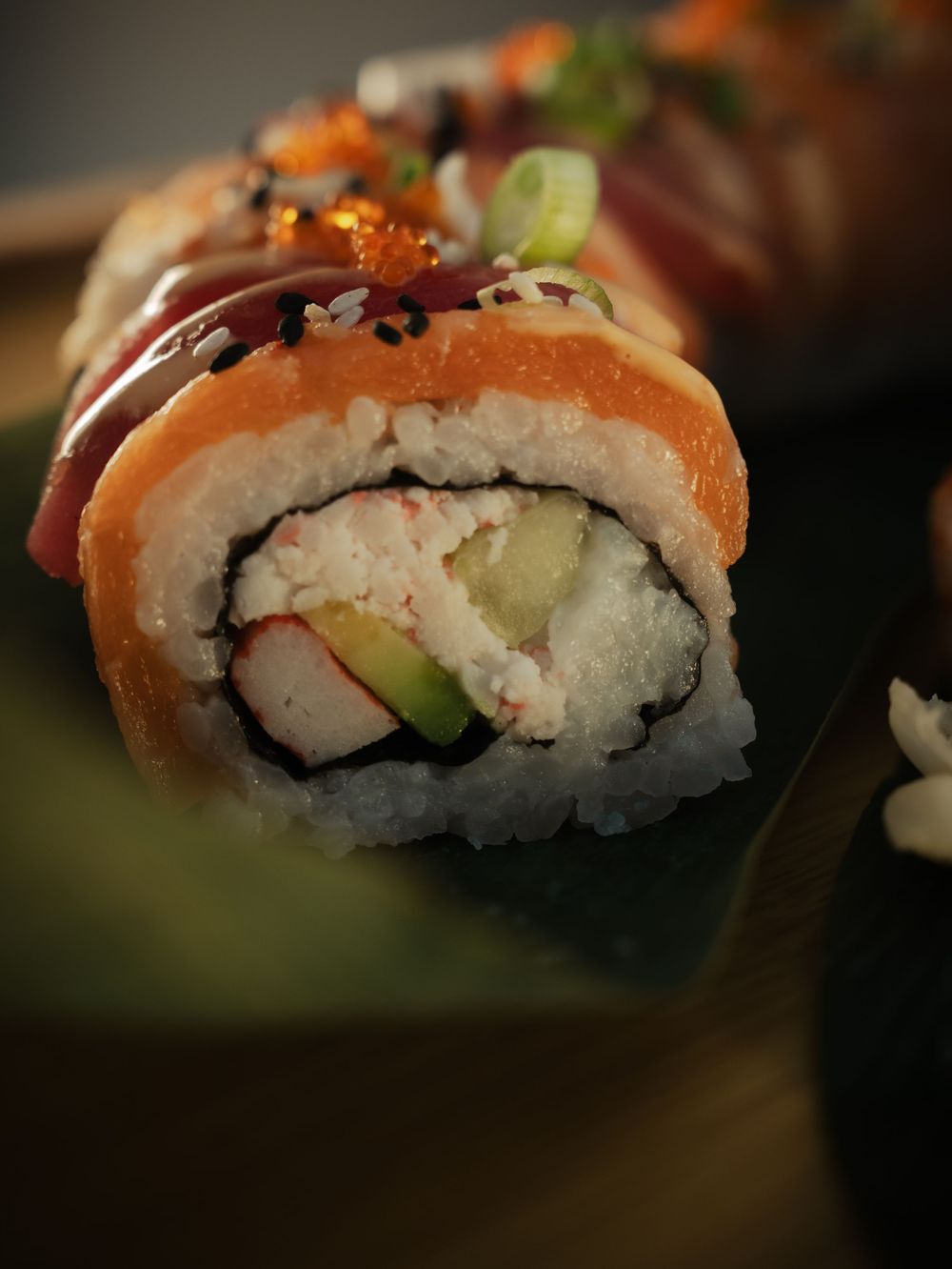 Close-up of a fresh salmon sushi roll with crab, avocado, and rice, topped with sesame seeds on a green leaf.
