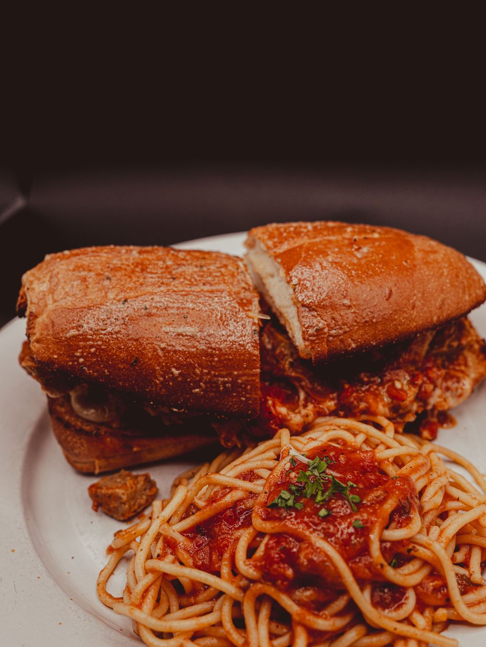 A plate with a saucy meatball sandwich on a brioche bun served alongside spaghetti in marinara sauce.