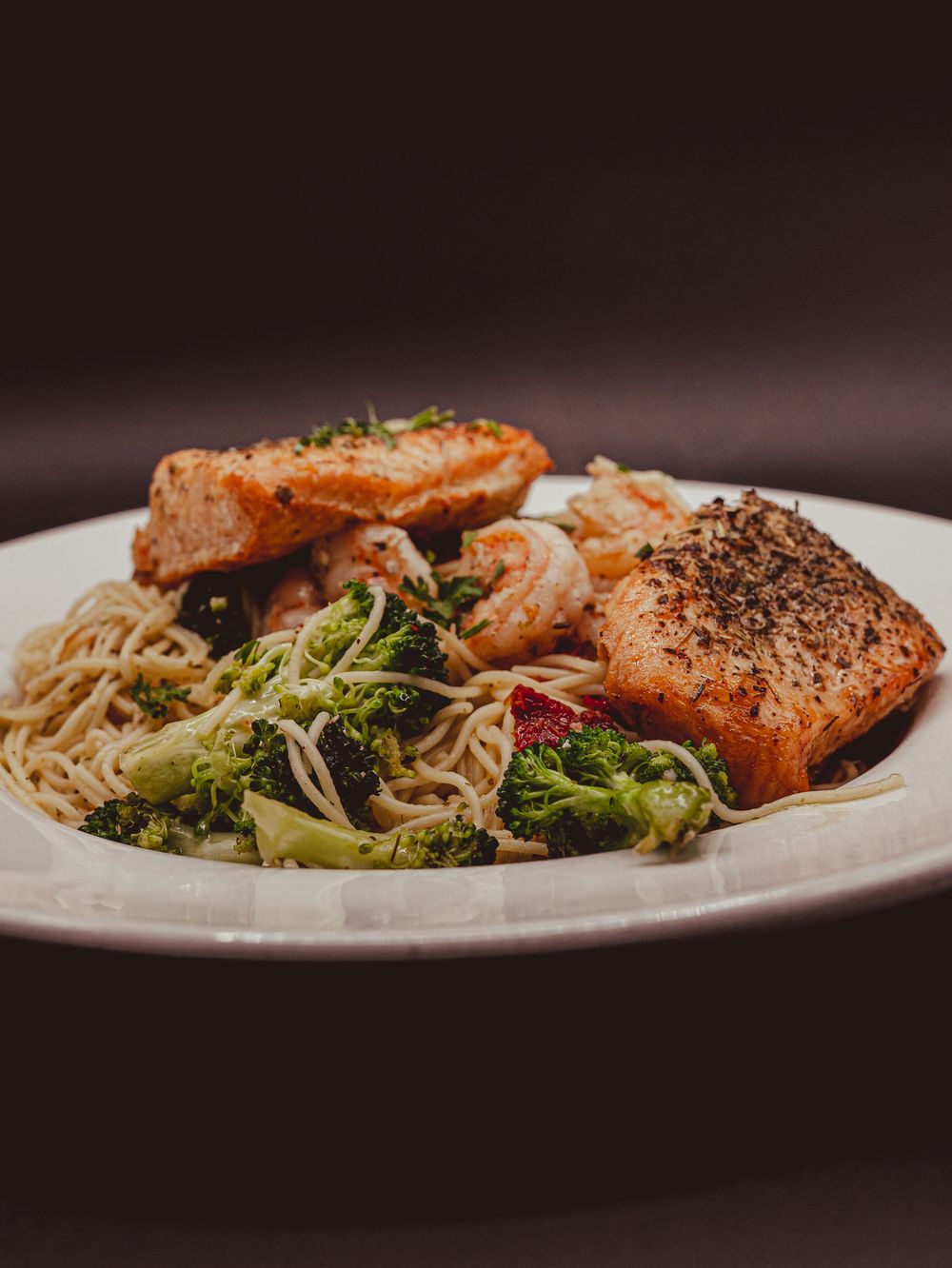 Grilled salmon fillets served over noodles with roasted broccoli and shrimp on a white plate.