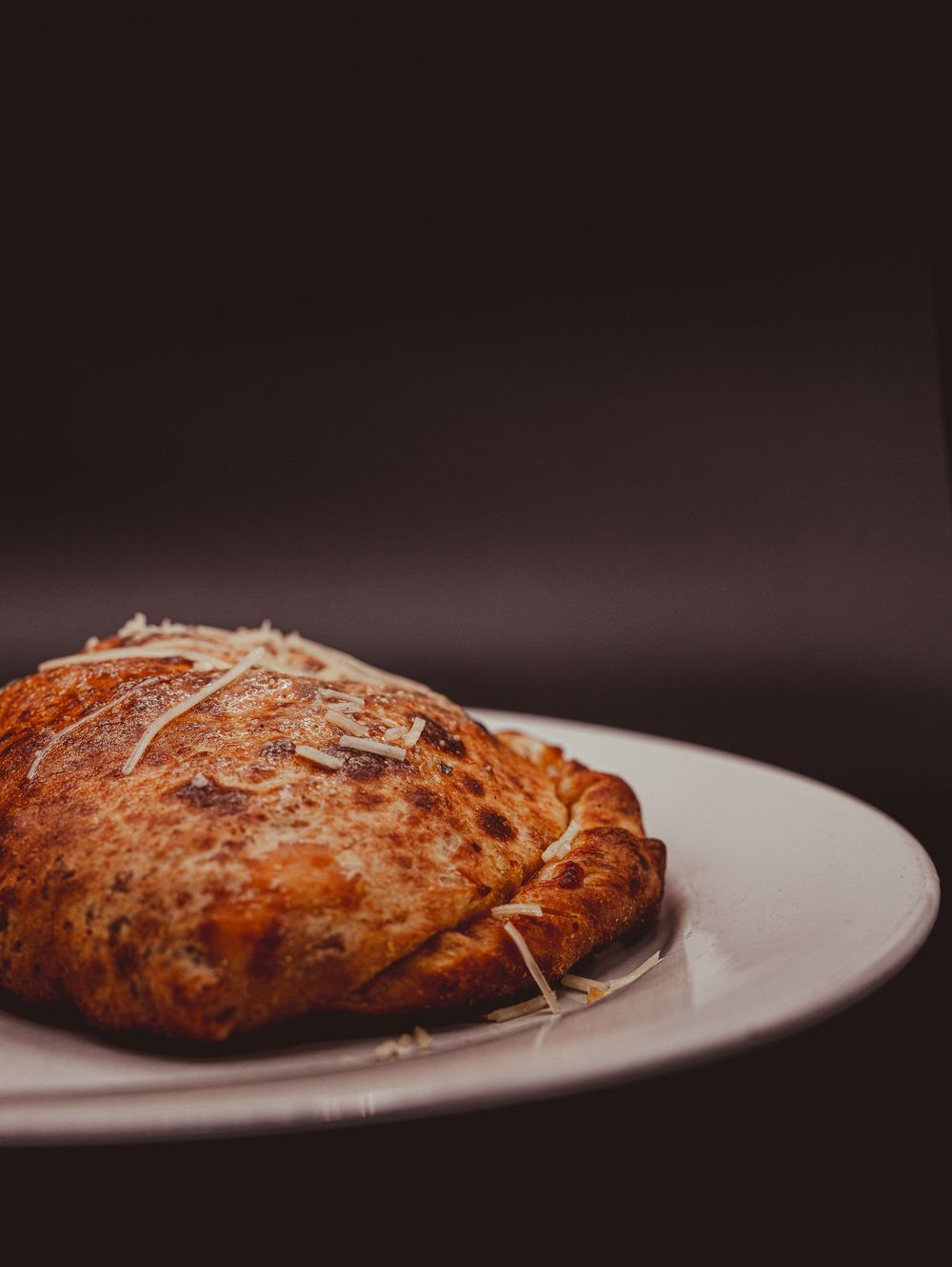 Golden-brown calzone topped with shredded parmesan cheese served on a white plate against dark background.