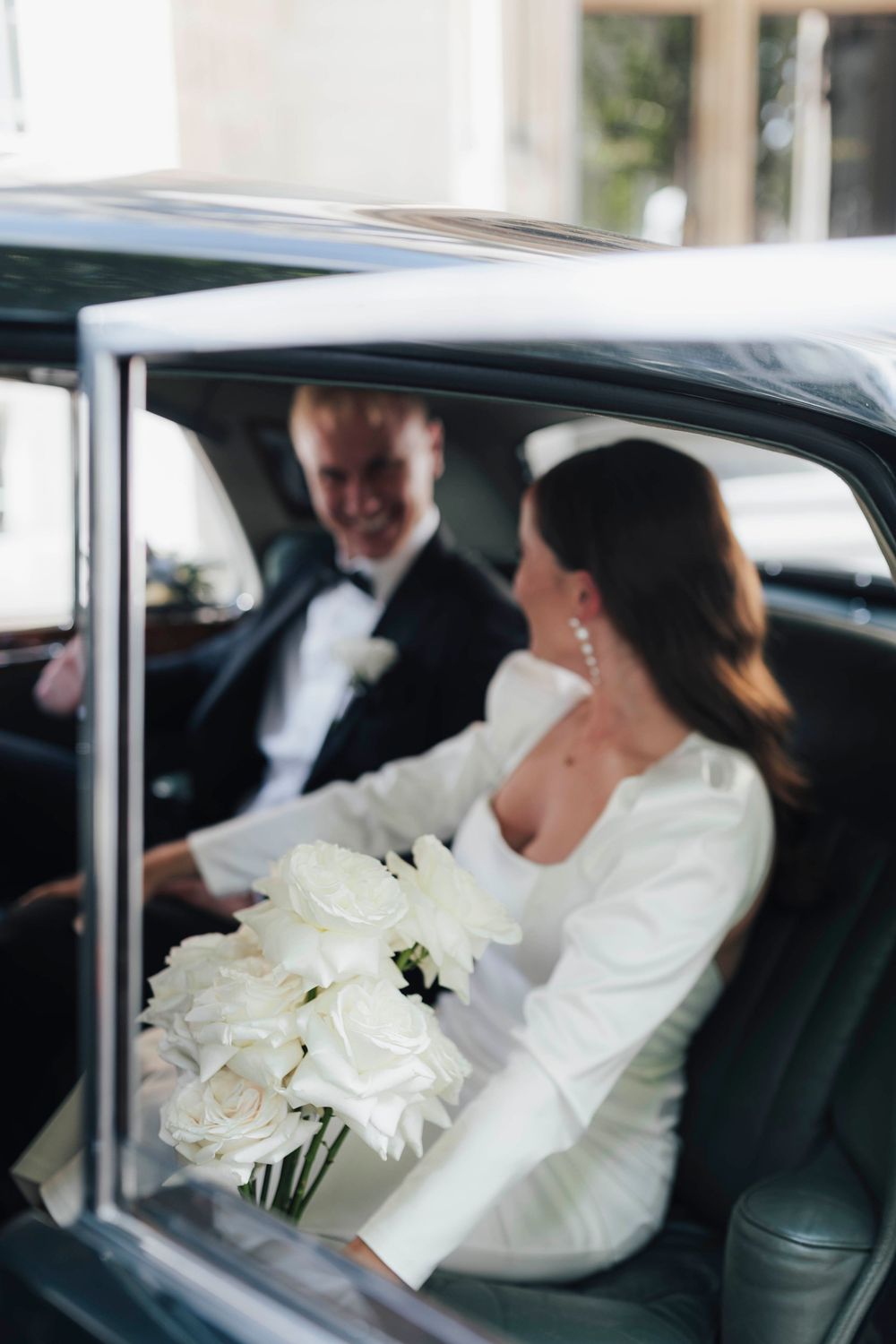 Bride and groom sitting in a vintage black car, bride holding a bouquet of white roses in an elegant wedding dress.