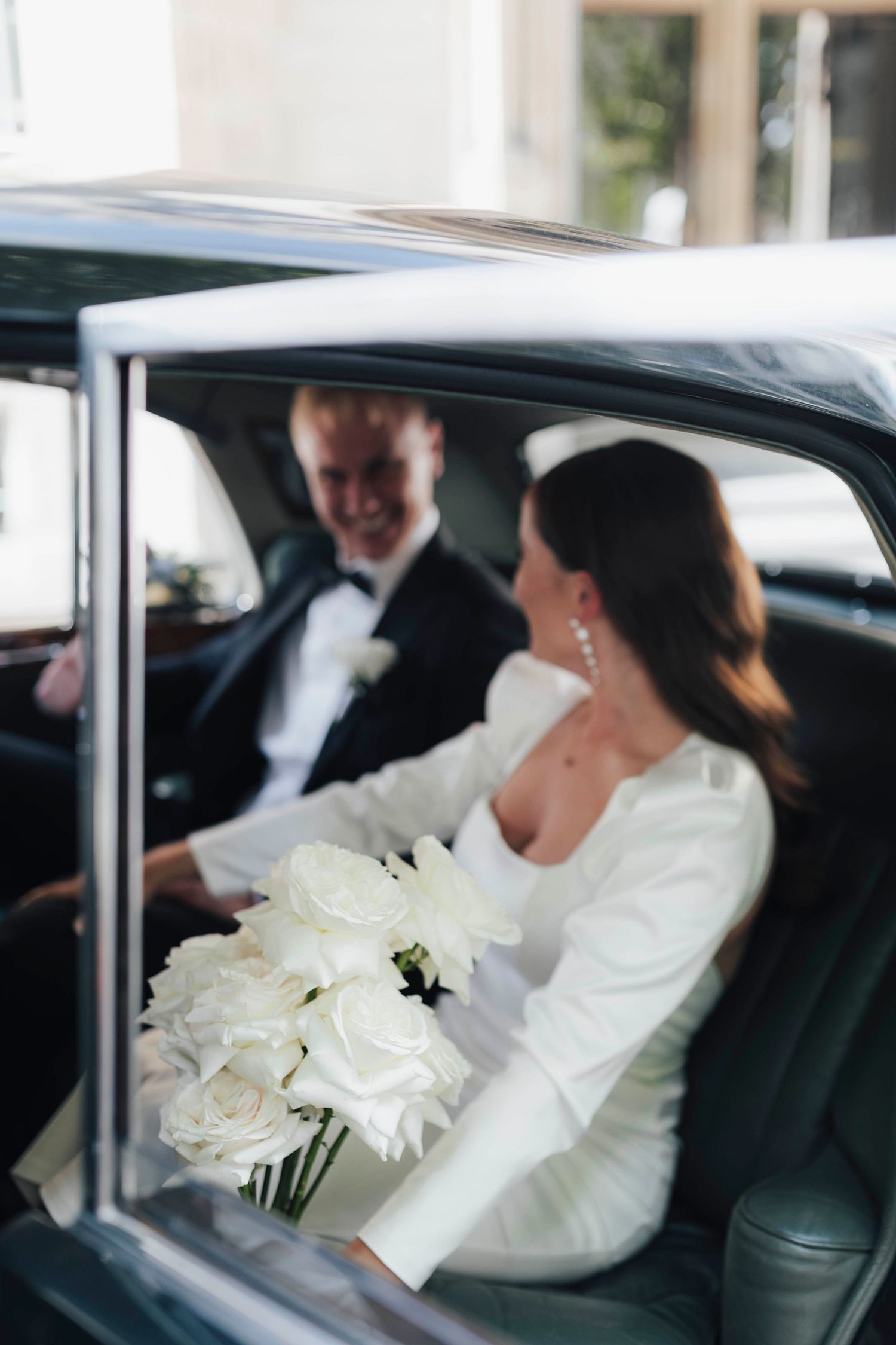 Bride and groom sitting in a vintage black car, bride holding a bouquet of white roses in an elegant wedding dress.