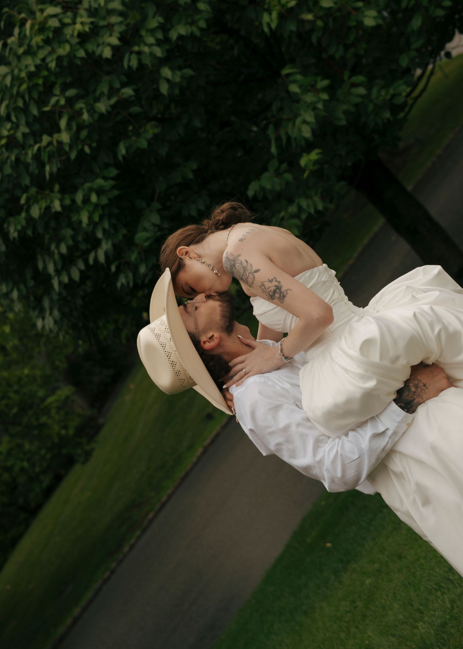A couple shares a romantic dip kiss outdoors, bride in white gown with tattoos, groom in white shirt and hat.