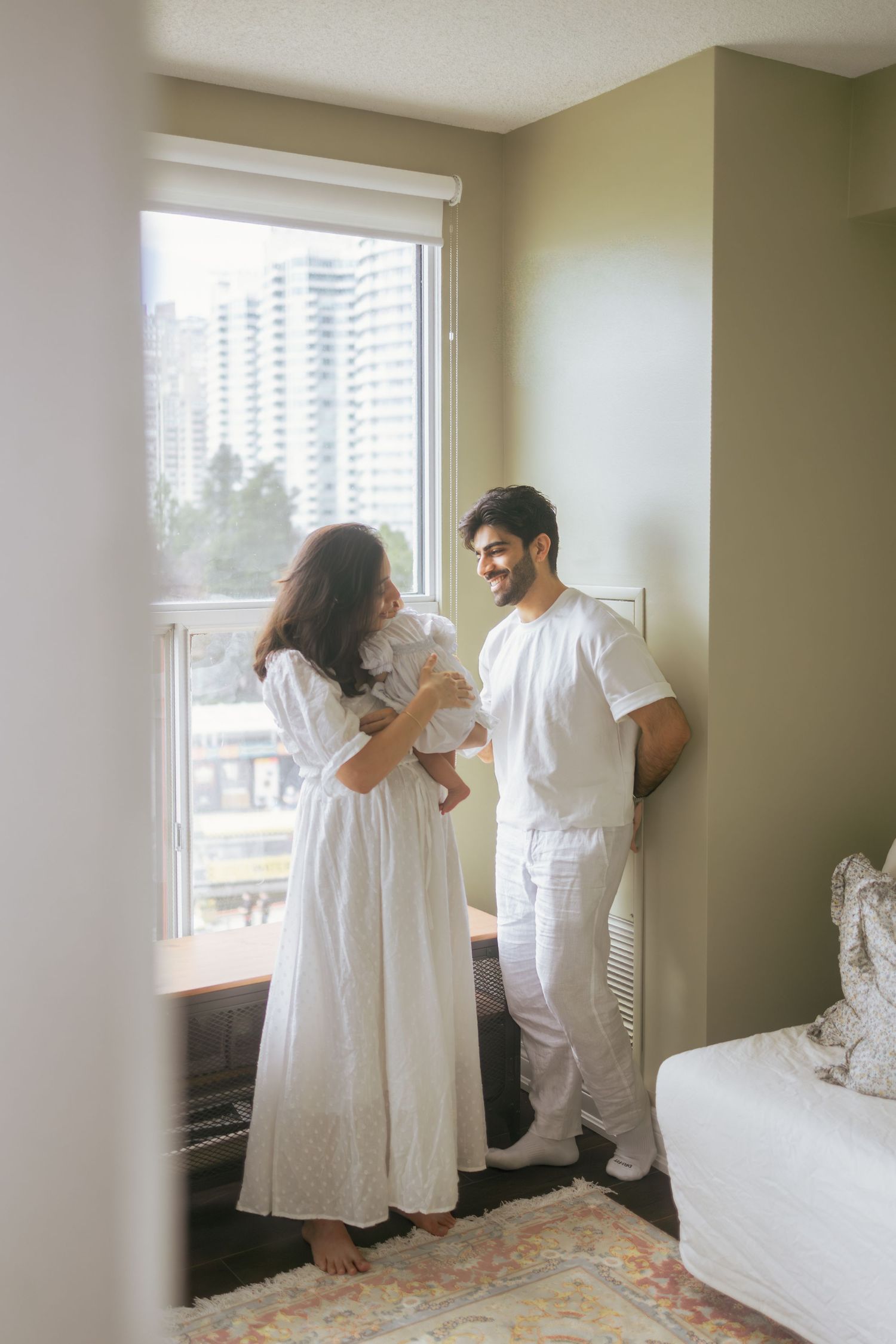 Young couple in white clothing sharing tender moment with baby near bright apartment window.
