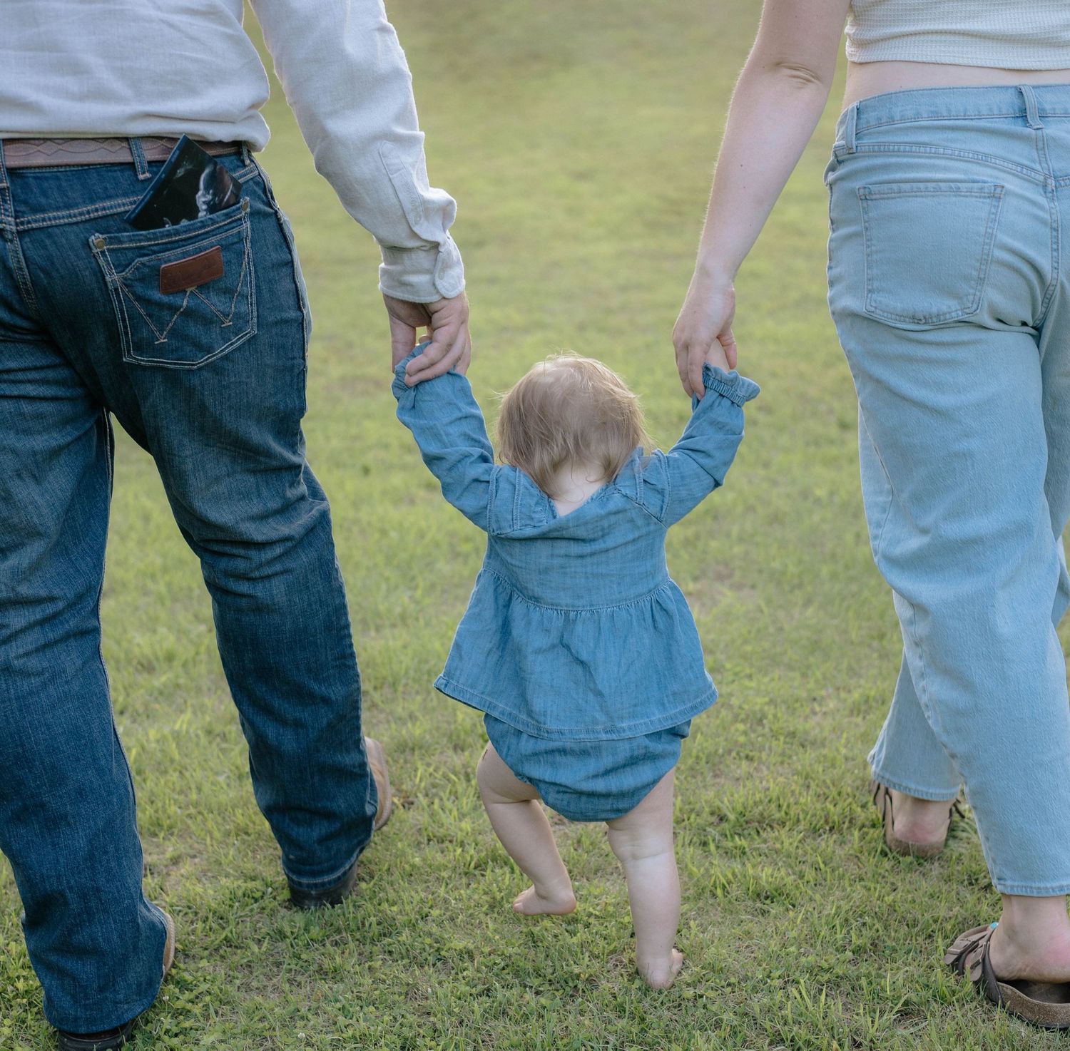 Baby in denim outfit holds parents hands while learning to walk on green grass outdoors.