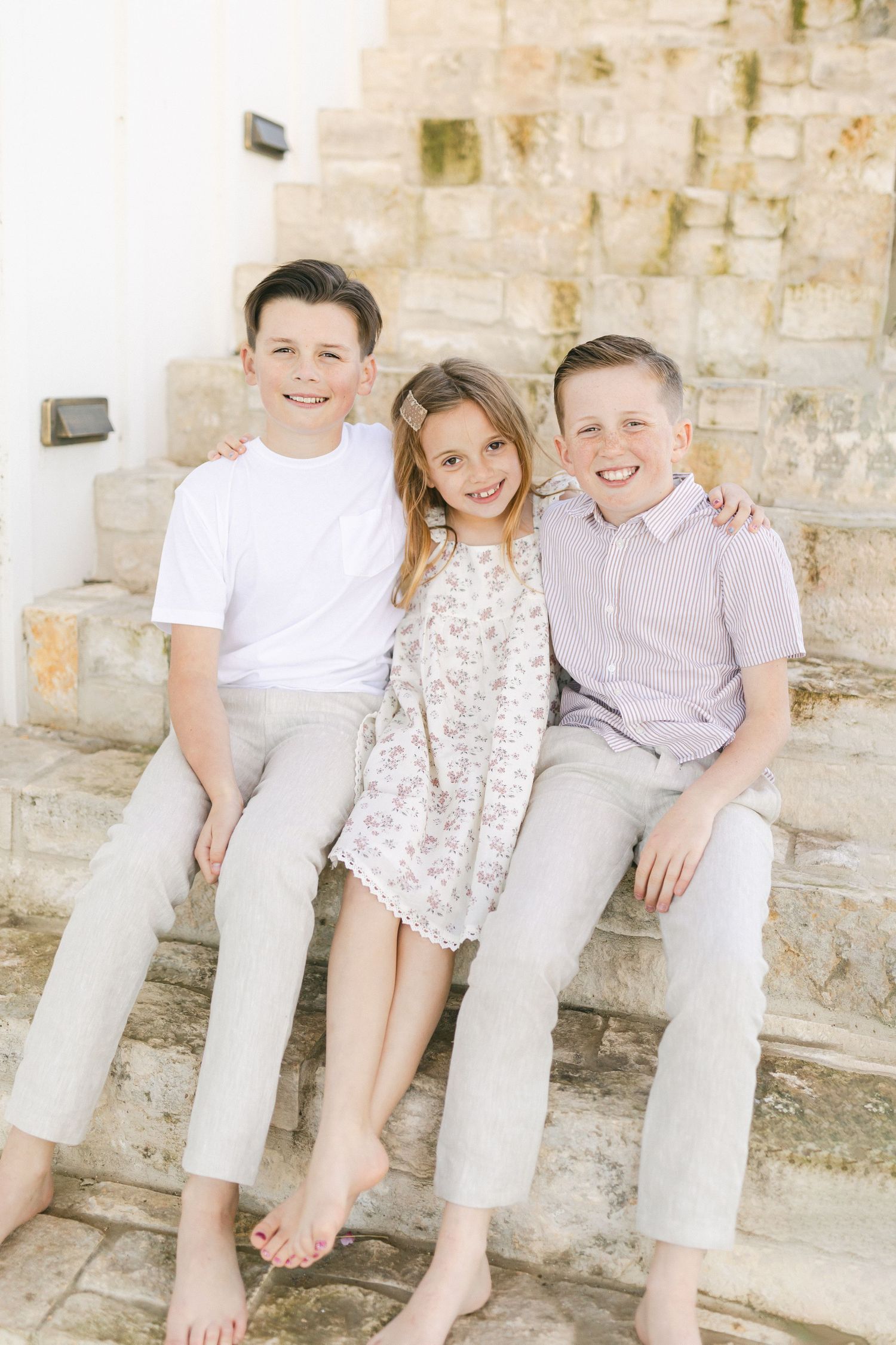 Three smiling children sitting together on stone steps, two boys in light clothing flanking a girl in a floral dress.