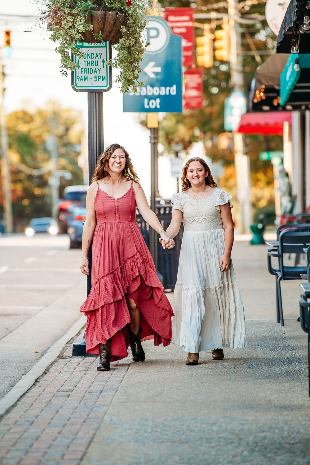 Two women in flowing dresses walk hand in hand along a sunny city sidewalk, smiling joyfully.