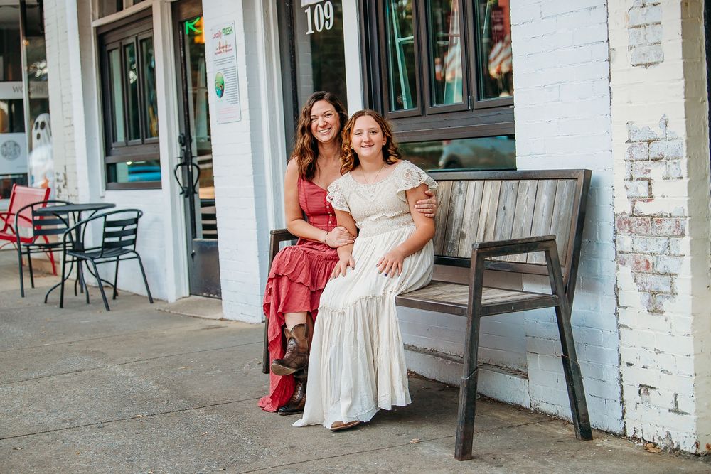 Two smiling women in dresses sit together on a bench outside a charming storefront.