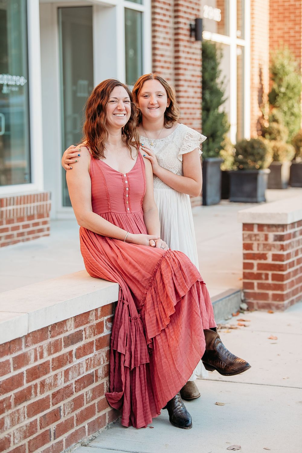 Two women in elegant dresses smile together while sitting on a brick ledge in an urban setting.