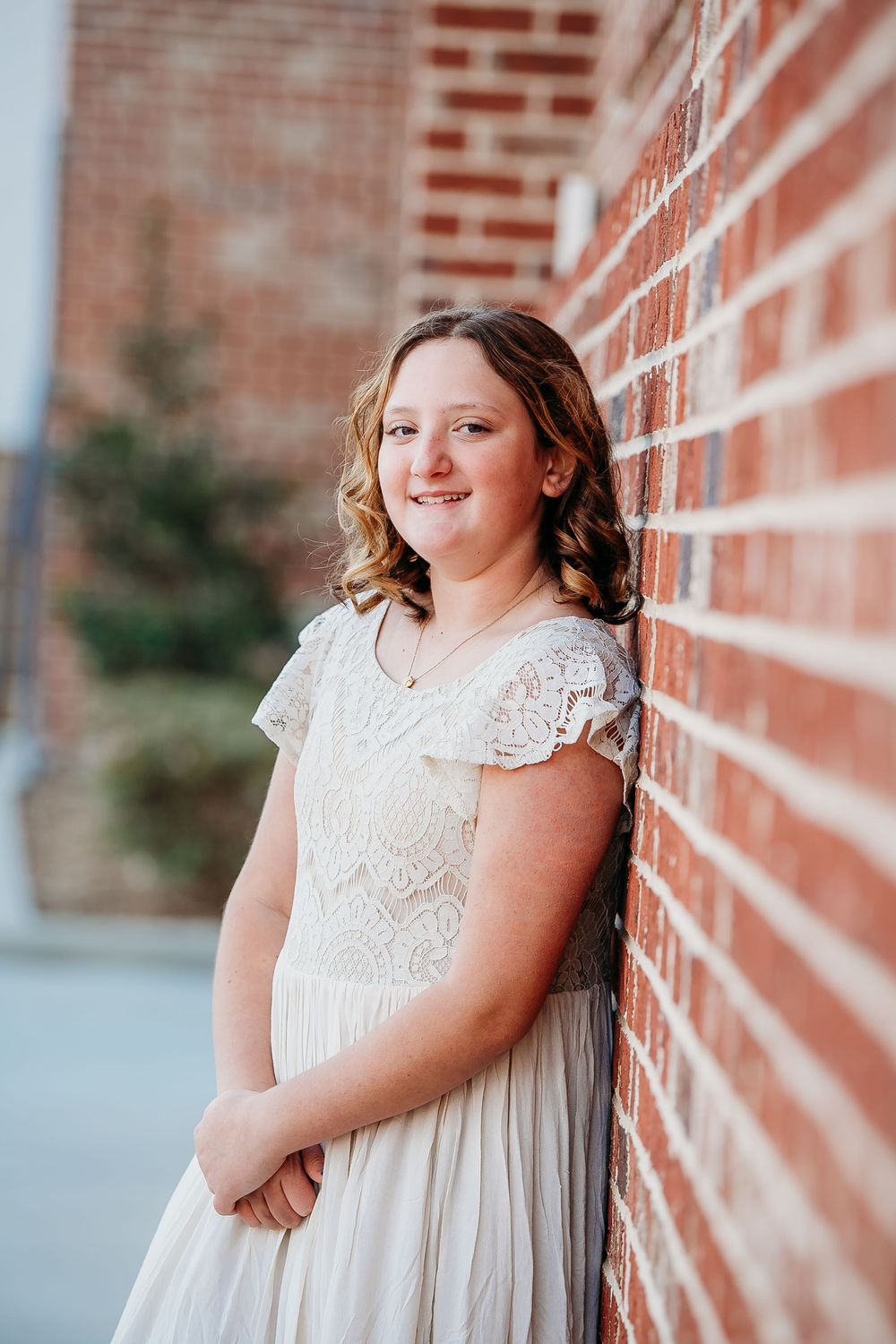 A young woman in a white lace dress leans against a brick wall, smiling softly outdoors.