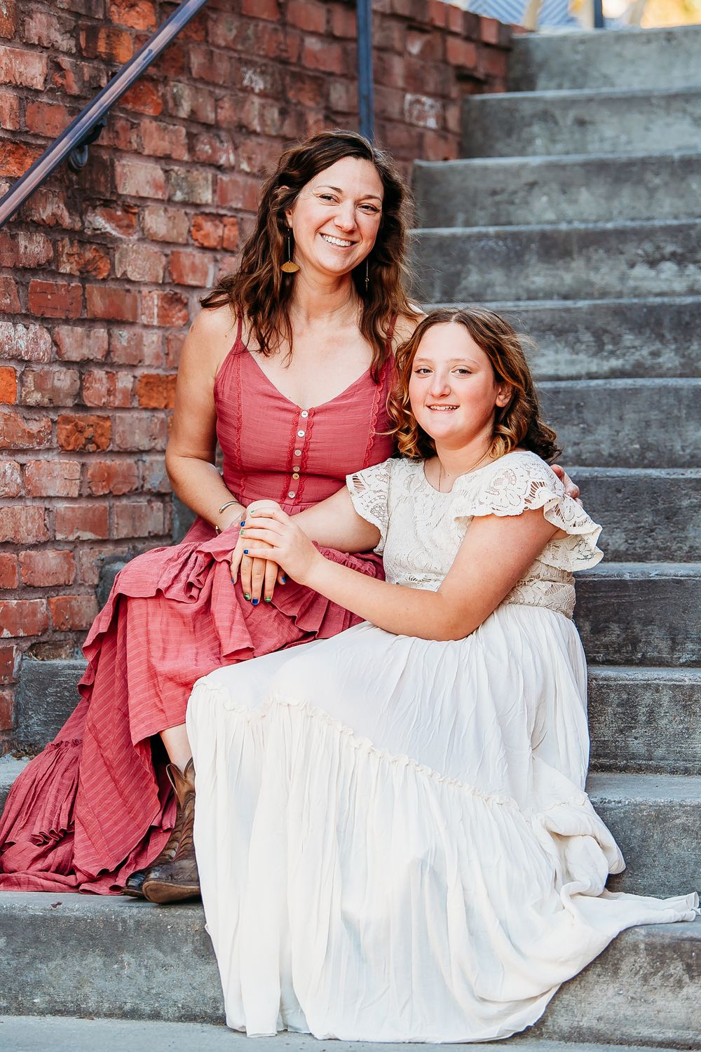 A smiling woman in a red dress and a girl in white sit together on brick steps.
