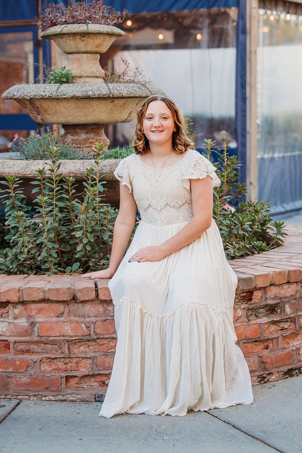 A young girl in a cream lace dress sits by a stone fountain outdoors.