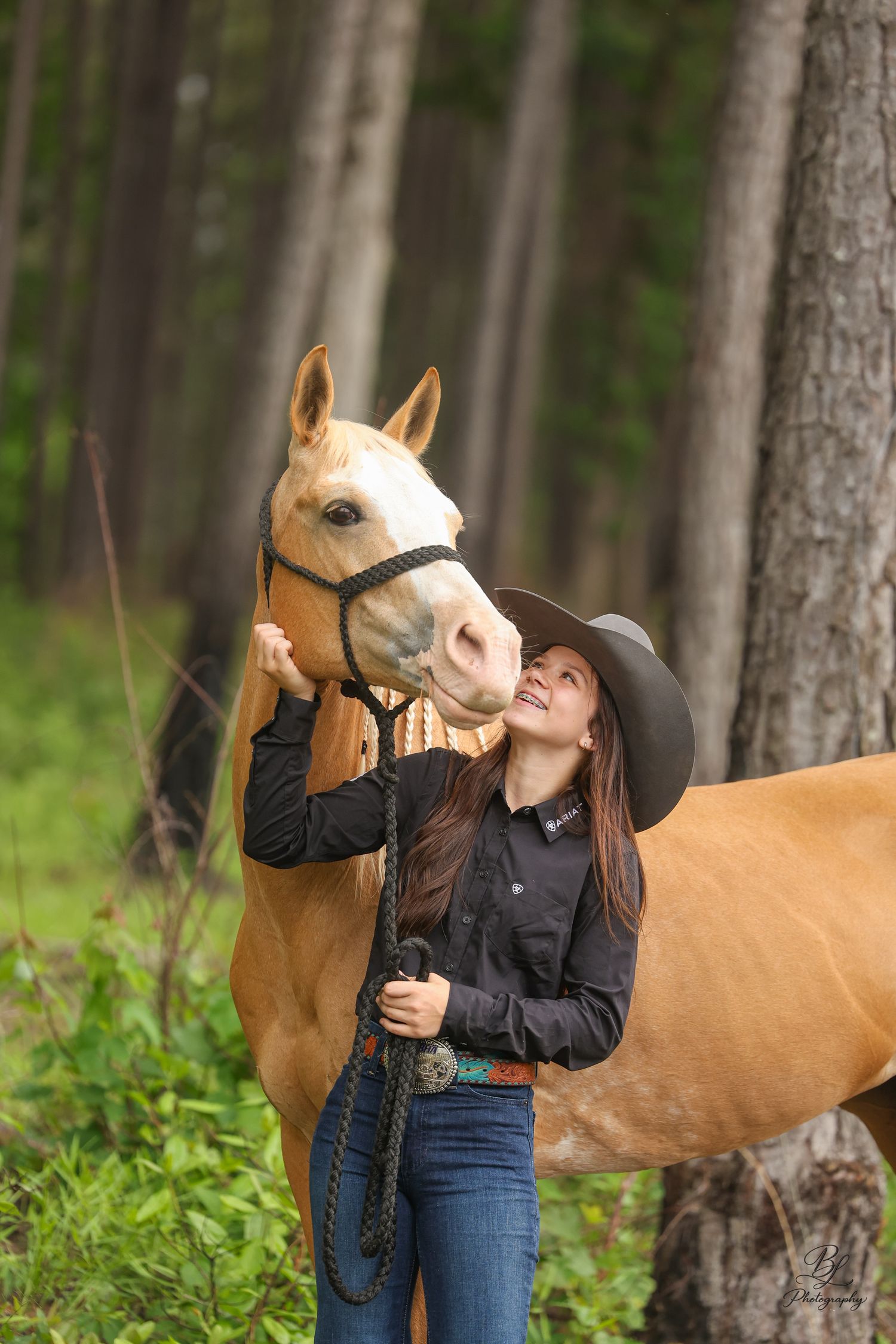 Young woman in cowboy hat smiling up at a golden palomino horse while holding reins in a forest setting.
