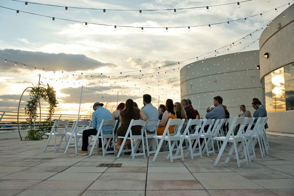 Outdoor rooftop wedding ceremony at sunset with white chairs, string lights, and floral arch.