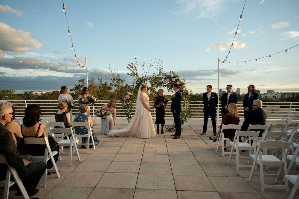Outdoor rooftop wedding ceremony with string lights, floral arch, bride and groom, officiant, and guests seated in white chairs at dusk.
